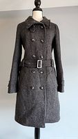 Front view of grey and black herringbone patterned coat on mannequin, showing double-breasted button closure and belted waist with buckle.
