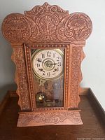 Full view showing carved wood gingerbread mantel clock with glass front panel and visible pendulum inside.