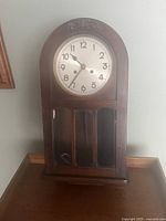 Front view of the wooden antique wall clock on a table, showing rounded top and clock face.