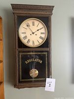 Full view of the vintage wooden regulator wall clock mounted on wall showing entire clock from top to bottom including clock face and pendulum case.