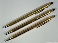 Three gold-tone Cross writing instruments displayed on white background: two ballpoint pens and one mechanical pencil. Top view showing black clickable tops and clips on two items.