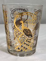 Close-up of a single rocks glass decorated with gold and black peacock motif on clear glass.