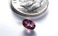 Oval faceted Rhodolite gemstone shown next to a 2024 dime for size comparison. The gemstone shows a deep pinkish-purple color with transparency and light reflection off facets.