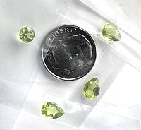 Four loose light green pear-shaped faceted peridot gemstones individually bagged and arranged around a U.S. dime for size comparison on a white background.