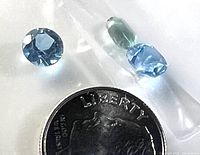Photo shows four loose Swiss Blue Topaz gemstones in various cuts and shades including a round one and a cushion cut, positioned beside a dime for scale.