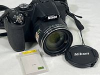 Front view of Nikon Coolpix P530 camera with attached lens, extra battery, and Nikon lens cap alongside.