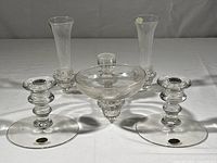 Set of two clear glass candleholders and a glass epergne centerpiece shown together. Items are clear glass, with wide bases and intricate stems. The epergne centerpiece has a broad dish part and a central cone-shaped component.