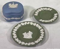 Top-down photo showing two circular sage green trinket dishes with white relief decoration and one blue rectangular trinket box with white relief designs.