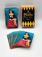 Photo showing complete deck of playing cards fanned out next to original box, card backs displaying pin-up artwork and box in fair condition with edge wear.