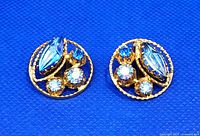 Front view of the pair of vintage gold tone clip-on earrings with blue and clear rhinestones against a blue textured background.