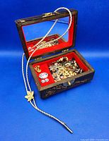 Asian themed carved wood jewelry box open showing red velvet interior with assorted jewelry inside including chains, bracelets, beads, clip-on earrings, and a long bolo tie extending outside the box.
