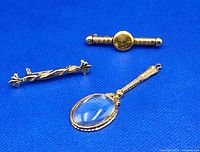 Top view of three gold tone jewelry pieces on blue background, showing magnifying glass pendant and two brooches.