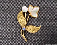 Close-up of vintage tulip brooch showing gold tone textured leaves, mother of pearl flower petal and bud, rhinestone detail, on dark textured background.
