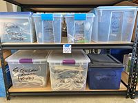 Two shelves filled with seven labeled plastic bins containing power strips, timers, floodlights, extension cords, and outdoor power strips. Bins are clear or blue with secure lids, handwritten labels visible.