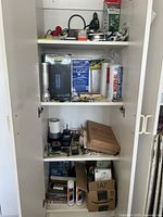 White shelving cabinet containing various tools, gadget boxes, and hardware items arranged on three shelves, including bug zapper, glue sticks, masking film, and other items.