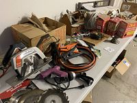 Photo of circular saw, jumper cables, boxes, and wiring on a white table.