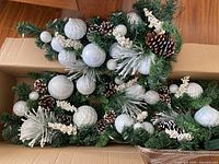 Overhead view of garland and matching centerpiece in a box showing white and frosted ornaments, pinecones, and white frosted berry sprigs with green foliage on wood floor.