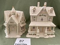 Two white Lenox ceramic Christmas Village houses with gold and green holly accents, one labeled Sweet Shoppe with turret and clock, side-by-side on green fabric backdrop.