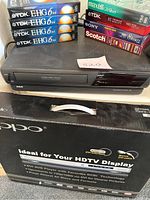 RCA VCR unit with multiple blank VHS tapes stacked on top and OPPO DVD player box in front