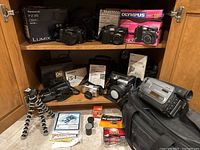 Multiple digital cameras including Panasonic Lumix FZ35, Olympus Camedia 3040 and C-770 Ultra Zoom, Olympus IS-1, Sony Handycam, original boxes, manuals, tripod and accessories displayed on shelves.