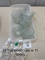 Top view showing 11 clear glass punch bowl cups stacked inside a plastic container alongside plastic bags containing metal hanging hooks.