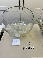 Top view showing large round glass bowl with 12 clear glasses nestled inside, placed on a table.