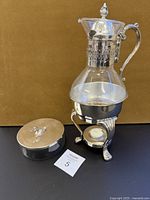 Photo showing glass coffee carafe with silver-plated ornate handle, lid, and warming stand. Lidded glass dish shown to the left.