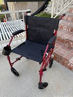 Front angled view of the red transport wheelchair showing lightweight frame, black fabric seat and backrest, right side cup holder attached to armrest, and projection of push handles and small front wheels.