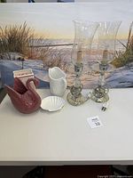 All items arranged on white surface in front of coastal beach canvas print background including swan planter, milk glass vase, shell dish, square ceramic container, and two hurricane lamps