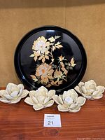 Full view of round black lacquered tray with gold floral decoration on wooden surface with ceramic flower decor underneath.