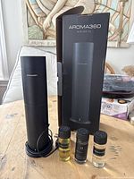 Aroma360 black scent diffuser device with three bottles of oils in front and original packaging box behind on wooden table.