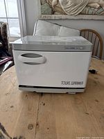 Front view of the compact white electric towel warmer on a wooden table showing handle and control panel.