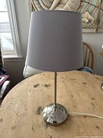 Full lamp showing metal base and gray fabric shade on wooden table near window.