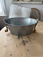 Oval galvanized steel bucket with rope handles placed on wooden table, showing side view and rope handles.