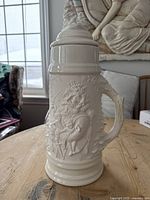 Front and side view of tall white ceramic stein on wooden table, showing raised relief of a bearded man playing a drum and pine tree lid with animal figure