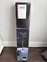 Box of artificial 9' light-up palm tree showing tree lit with string lights, indication of height and brand 'Canvas'.