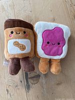 Peanut butter and jelly plush toys close together on wood surface, showing front face details and legs.