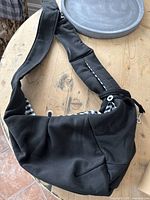 Full view of black fabric pet sling with checkered lining and metal clip closure shown on wooden table.