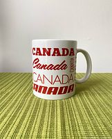 Photo of white ceramic mug showing 'Canada' text and handle on green surface