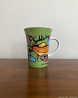Side view of ceramic mug with colorful Indigenous art on green background, showing handle and design details