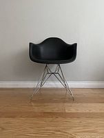 Front view of modern black plastic armchair with chrome metal wire base against plain white wall and wooden floor.