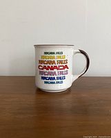 Front view of the white ceramic mug with Niagara Falls and Canada text in multiple colors, brown rim and handle visible.