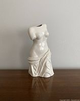 Front view of white ceramic vase shaped as a nude female torso with draped fabric on lower half.