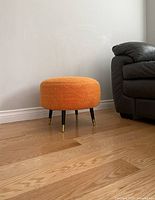 Full view of an orange footstool showing the textured fabric surface and four tapered black legs with brass caps at bottom next to a leather couch.