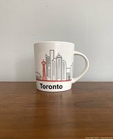 Front view of white ceramic mug displaying Toronto skyline artwork and text.