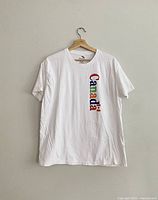 White short-sleeve t-shirt on a wooden hanger with 'Canada' text vertically printed in varied colors and a small Canadian flag