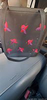 Black handbag on vehicle seat showing front with multiple red embroidered accents shaped like birds or flowers. Thin black handles visible.