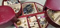 Open red circular jewelry case revealing multiple beige felt-lined sections with assorted fashion jewelry such as rings, earrings, bracelets, and small jewelry supplies.