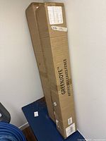 Two long cardboard boxes leaning against a wall, labeled Greenjoye Artificial Leaves Fence.