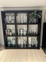Front view of black IKEA shelf with glass and glassware inside compartments to show scale and condition.